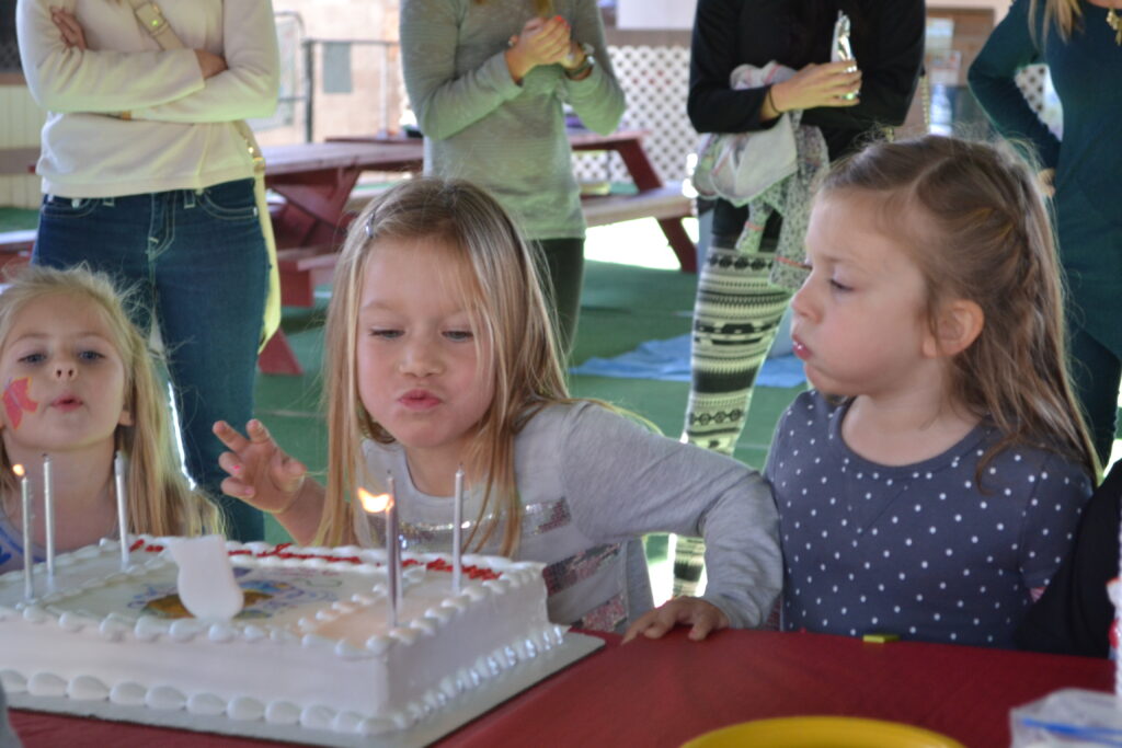 Three elementary school age girls blowout the candles on a large white sheet cake while four adults stand behind them.