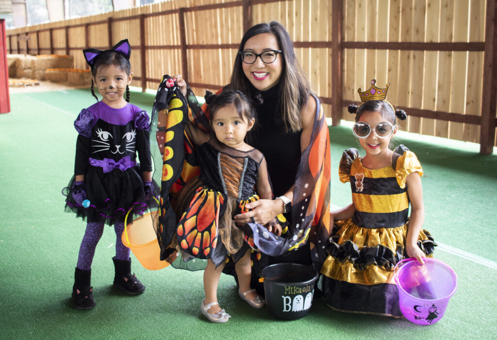 Three little girls and their Mom, with their buckets of candy, are all dressed up for Halloween as a black and purple cat, an orange and black butterfly and a princess with a yellow dress.