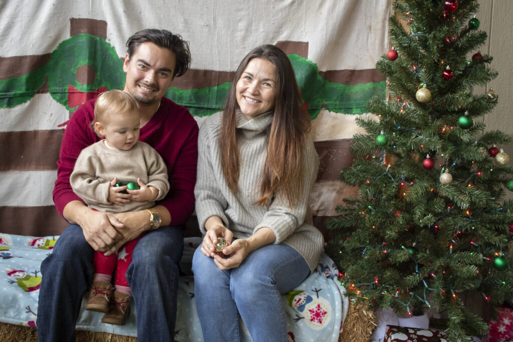 A man in a red sweater and a woman in a grey turtleneck sweater sitting next to a Christmas tree while holding their toddler aged son.